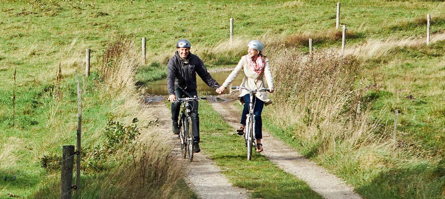 An einem Frühlingstag radeln zwei Personen auf einer Radtour entlang des Hærvejen bei Gudenåens Udspring Hand in Hand