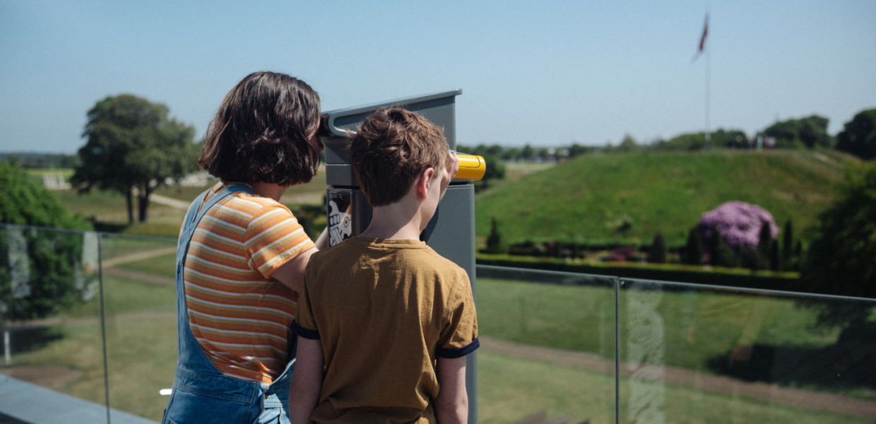 Zwei Kinder blicken durch ein Fernglas von der Dachterrasse von Kongernes Jelling auf die Hügel